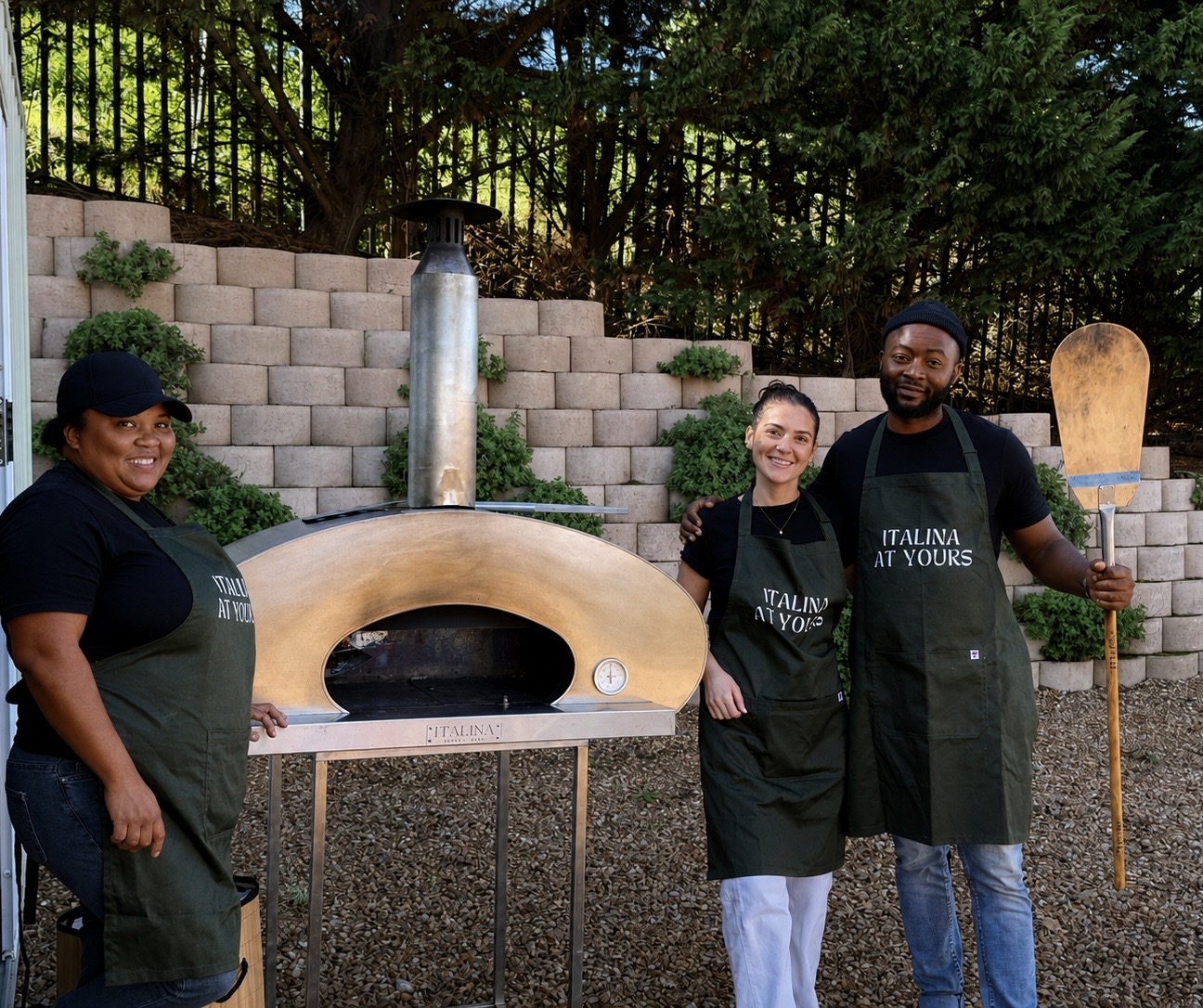 The Italina At Yours crew in branded aprons beside the wood-fired oven with a pizza peel