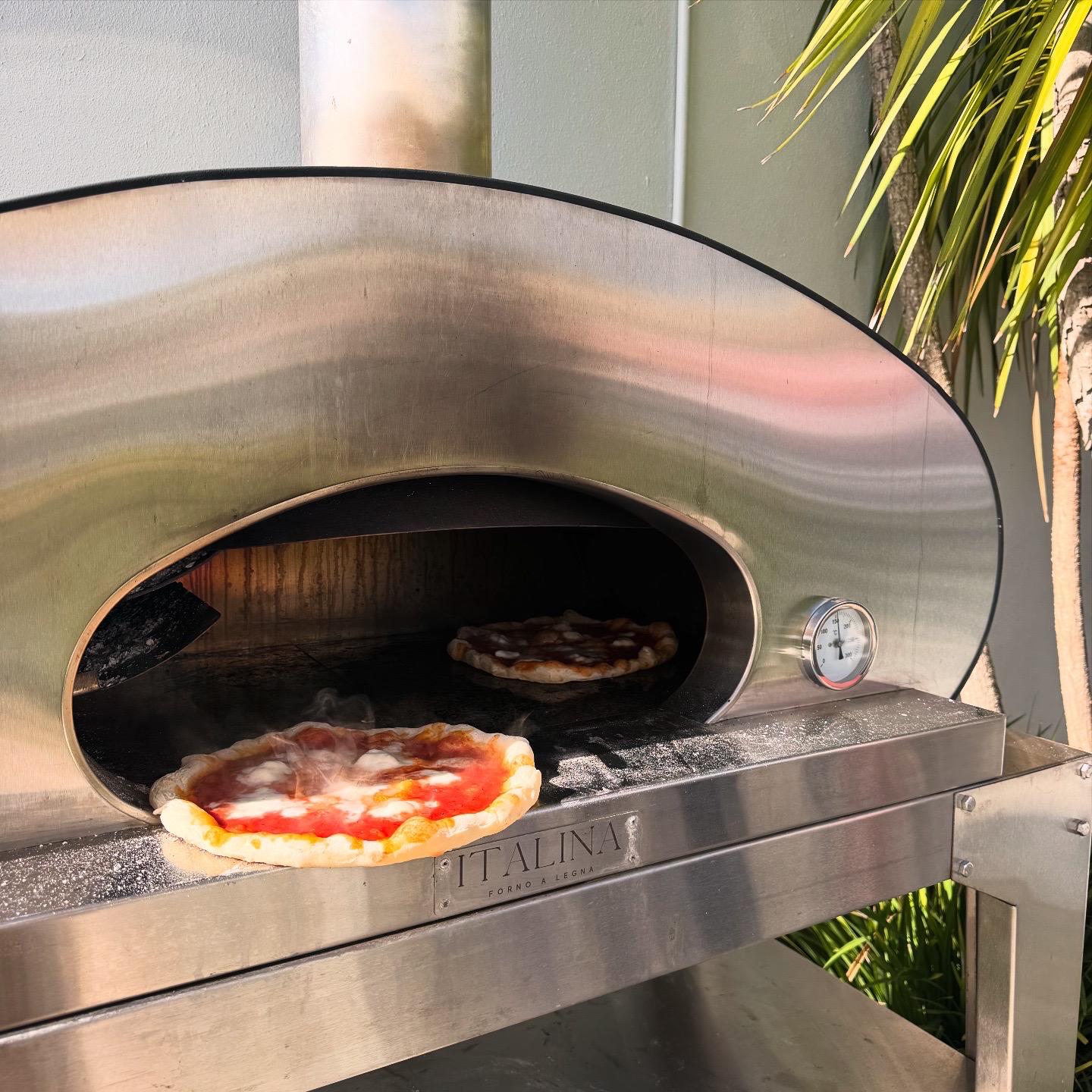 Two freshly made pizzas cooking in the wood-fired oven beside a palm